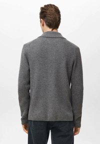 Szary sweter z dzianiny o prążkowanej fakturze, z długimi rękawami i kołnierzem typu szalowego, noszony z ciemnymi spodniami, widziany od tyłu.