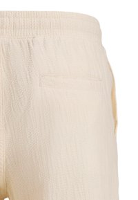 Pantalon beige clair texturé avec une ceinture élastique et une seule poche arrière, présentant un motif diagonal subtil sur le tissu.