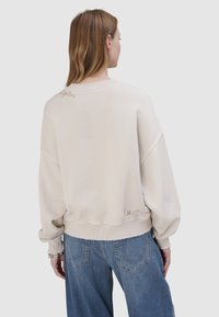 Kobieta o brązowych włosach, ubrana w luźną, białawą bluzę z drobnym napisem na plecach i niebieskie dżinsy, stojąca na tle jednolitego szarego tła.