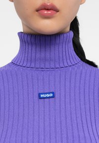 Maglione a collo alto a coste viola con un patch blu "HUGO" sul davanti. Il materiale ha una consistenza morbida e un design aderente.