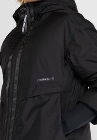 Chaqueta impermeable negra con cremallera frontal, capucha ajustable y tejido texturizado. Presenta un logo en el pecho y puños con ojal para el pulgar.