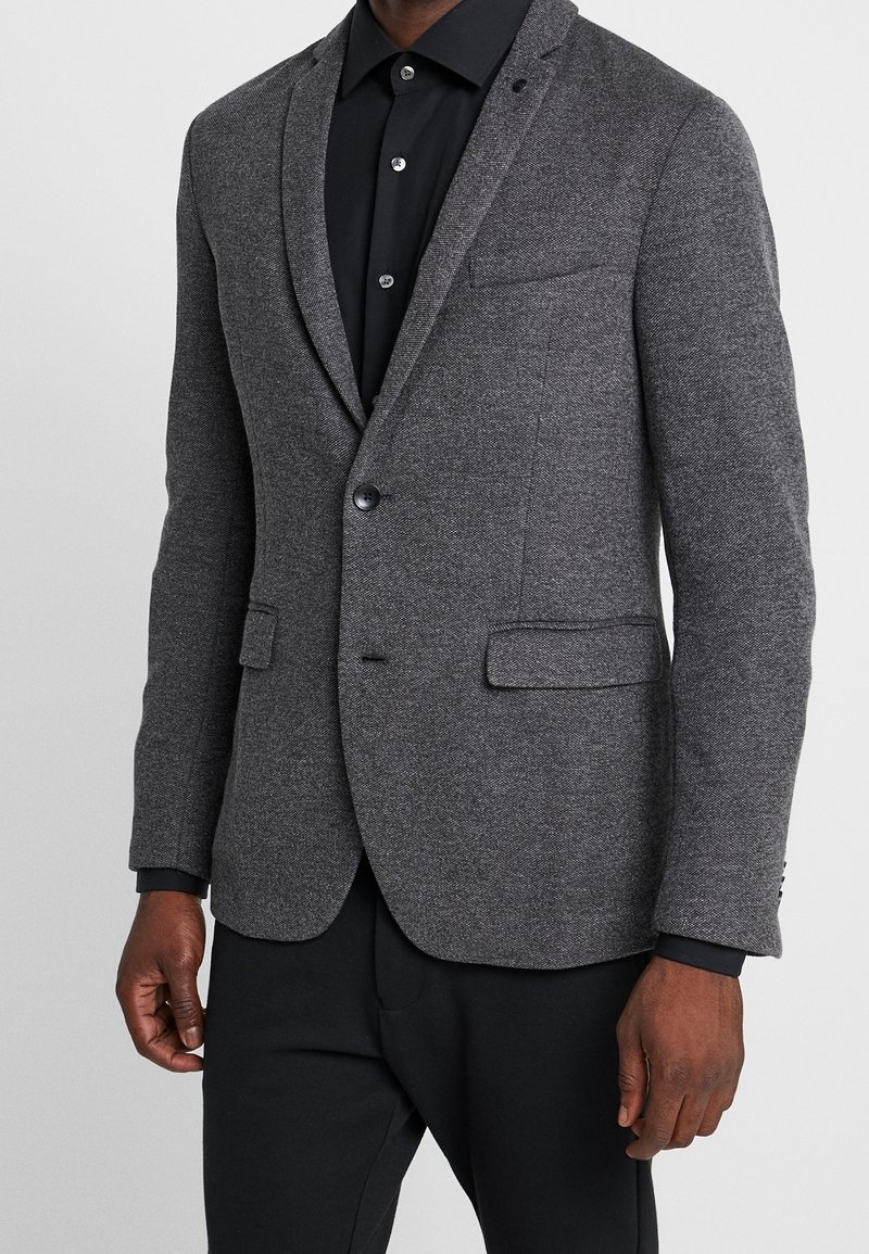 Blazer de mezcla de lana gris con solapa de muesca, cierre de un botón y dos bolsillos frontales. Estilizado sobre una camisa negra y combinado con pantalones negros.