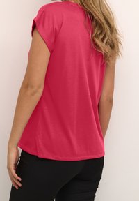 Maglietta a maniche corte in tessuto rosa brillante, con una vestibilità ampia, orlo curvo e spalle leggermente scese, abbinata a pantaloni neri.