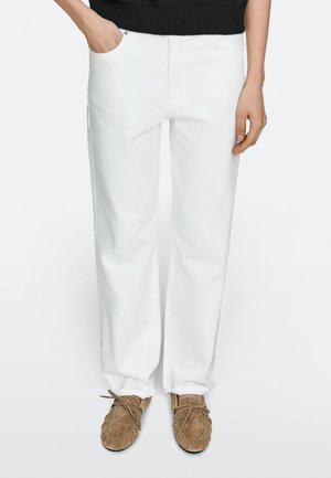 Person trägt weiße, gerade geschnittene Hosen und braune Wildleder-Mokassins, eine Hand in der Tasche, steht vor einfarbigem Hintergrund.