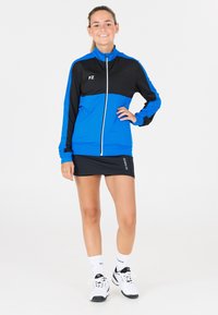 Blå och svart idrottsjacka med hel dragkedja och hög krage, kombinerad med en svart kjol och vita sportskor. Mjuk och lätt vikt.