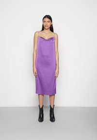 Vestido de seda roxa com decote drapeado e alças finas, combinado com botas de tornozelo pretas com bico fino, sobre um fundo cinza-claro.