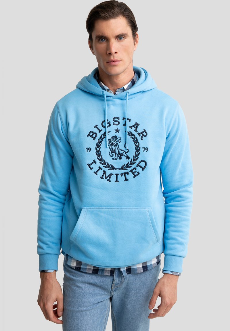 Světle modrá mikina s kapucí a přední kapsou, s černým grafickým logem, šňůrkovou kapucí a žebrovanými manžetami. Nositá přes károvanou košili.