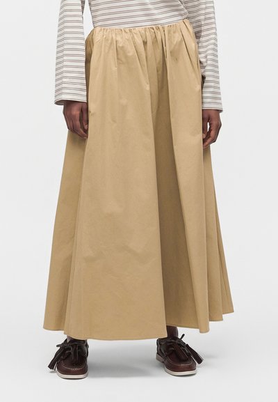 Jupe beige longueur cheville avec taille froncée portée avec des chaussures marron à pampilles et une chemise blanche à manches longues avec de fines rayures horizontales.