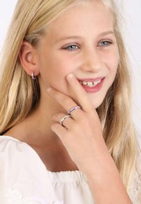 Zilveren en paarse ringen aan een hand, gecombineerd met kleine oorbellen. Blond haar en blauwe ogen, gekleed in een witte blouse. Subtiele textuur.
