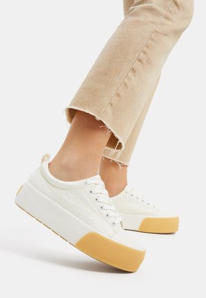 Füße in weißen Plateausneakern mit gelben Zehenkappen und ausgefransten, beigen, kurzen Hosen vor weißem Hintergrund.