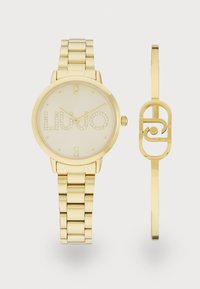Gold-tone watch with a round face, cream dial, studded numerals, and a linked bracelet. Accompanied by a matching gold cuff bracelet.