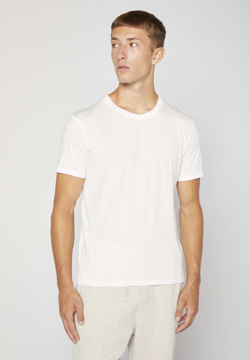 Vit kortärmad t-shirt med rund hals, lätt tyg, slät struktur och enkel design, bärs tillsammans med grå sweatpants.