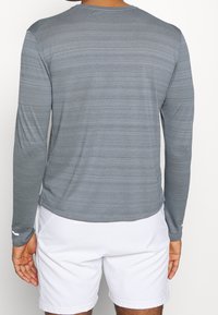 Langärmliges graues Performance-Shirt mit horizontalen Streifen, Rundhalsausschnitt und weicher Textur. Dazu gibt es weiße Shorts.