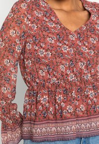 Blusa floreale rossa con scollatura a V, vita elastica e maniche lunghe con volant. Presenta un orlo decorativo con una banda a fantasia contrastante.