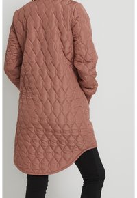 Une personne portant une longue veste matelassée couleur rose poussiéreux avec un motif en losanges, associée à un pantalon noir, se tenant devant un fond clair.