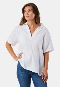 Weiße Bluse mit kurzen Ärmeln, V-Ausschnitt und lockerer Passform. Texturiertes Material mit einem subtilen Kräuselmuster. Der Saum ist an den Seiten etwas länger.