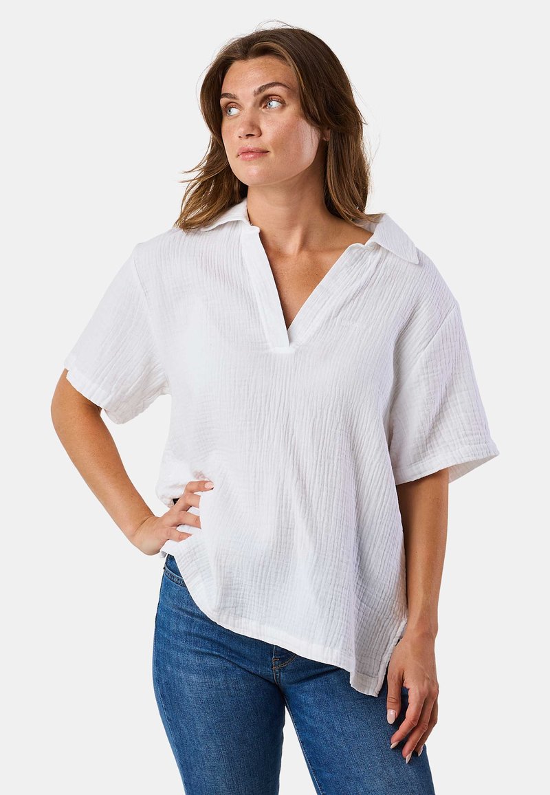 Weiße Bluse mit kurzen Ärmeln, V-Ausschnitt und lockerer Passform. Texturiertes Material mit einem subtilen Kräuselmuster. Der Saum ist an den Seiten etwas länger.