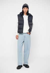 Námornícky prešívaný vest s bielou mikinou, skombinovaný s svetlomodrými džínsami. Čierne topánky a vzorovaná pletená čiapka dotvárajú vzhľad.