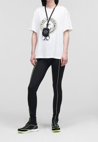 Witte oversized T-shirt met grafische print, zwarte legging met groene zijkanten, en zwarte sneakers met groene accenten en ritssluiting.