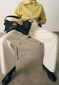 Pull jaune tricoté à fermeture éclair, pantalon large blanc, sac à main en cuir noir avec ferrures dorées, associé à des bottines à talons noires.
