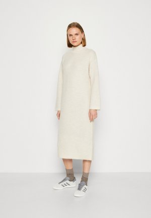 Rochie tricotată crem cu guler înalt, mâneci lungi și o siluetă dreaptă, asortată cu adidași albi și șosete maro cu model în combinație.