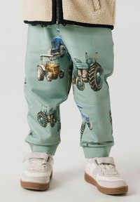 Pantaloni della tuta verdi con una stampa di trattori in vari colori, dotati di polsini elastici alle caviglie, abbinati a sneakers bianche e grigie.