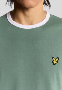 Persona che indossa una maglietta verde girocollo con bordi bianchi e un piccolo logo di un uccellino giallo ricamato sul lato sinistro del petto.