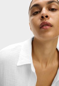Camicia bianca strutturata con colletto, caratterizzata da un sottile motivo sgualcito. Modello con pelle chiara mostrato in un'inquadratura ravvicinata.