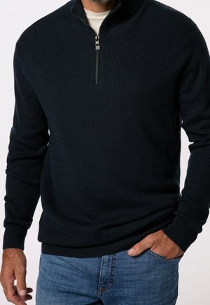 Homme portant un pull côtelé bleu marine foncé à demi-fermeture éclair sur une chemise blanche et un jean bleu, avec une main dans la poche et l'autre détendue sur le côté.