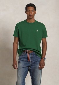 Jeune homme portant une chemise verte à manches courtes avec un petit logo, un jean bleu, une ceinture en tissu rayée et une montre-bracelet, se tenant devant un fond uni.