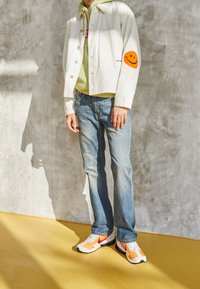 Veste blanche courte avec patch smiley orange, sweat à capuche vert, jean bleu clair et baskets blanches avec accents oranges sur un sol jaune.
