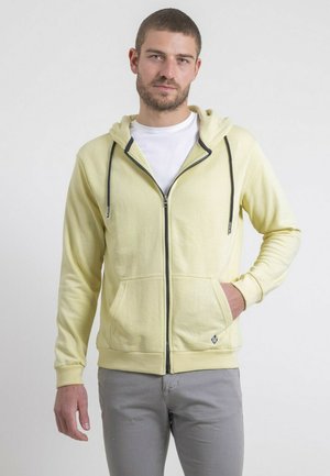 Homme aux cheveux courts et barbe, portant un sweat à capuche zippé jaune pâle et un pantalon gris, debout avec une main dans la poche.