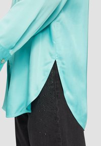 Een turquoise satijnen overhemd met een high-low zoom, gecombineerd met zwarte pantalons met strassaccenten bij de zakken.