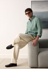 Camicia a maniche lunghe verde chiaro, pantaloni beige e mocassini neri. Modello seduto su un divano grigio imbottito, in posa con una gamba sull'altra.