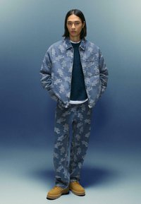 Tânărul poartă o jachetă din denim cu model albastru �și pantaloni asortați, un pulover închis la culoare, o cămașă albă și pantofi bej cu șireturi.