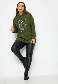 Olivgrön hoodie med stor grafisk tryck, tillsammans med svarta byxor i konstläder och svarta snörskor, vilket skapar en avslappnad outfit.