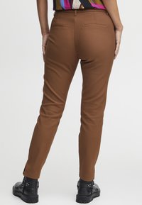 Hnědé slim-fit kalhoty s hladkou texturou, s kapsami na zadní straně a klasickým pásem. Nosené s černými kotníkovými botami.