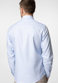 Jasnoniebieska koszula z długim rękawem, z kołnierzykiem zapinanym na guziki, gładki materiał, plisowanie z tyłu, zaokrąglony dół oraz mankiety zapinane na guziki. Prosty, klasyczny wzór.