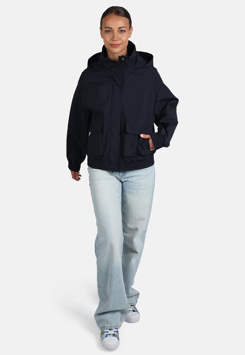 Granatowa wodoodporna kurtka z kapturem, dwoma przednimi kieszeniami, elastycznymi mankietami i regulowaną talią, w zestawie z jasnymi dżinsami i trampkami.
