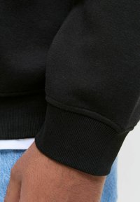Mustra musta särk, mille käisel on soonikkoes ja õmbluste detail. Close-up vaade näitab kanga sujuvust ja venitatud sobivust randme ümber.