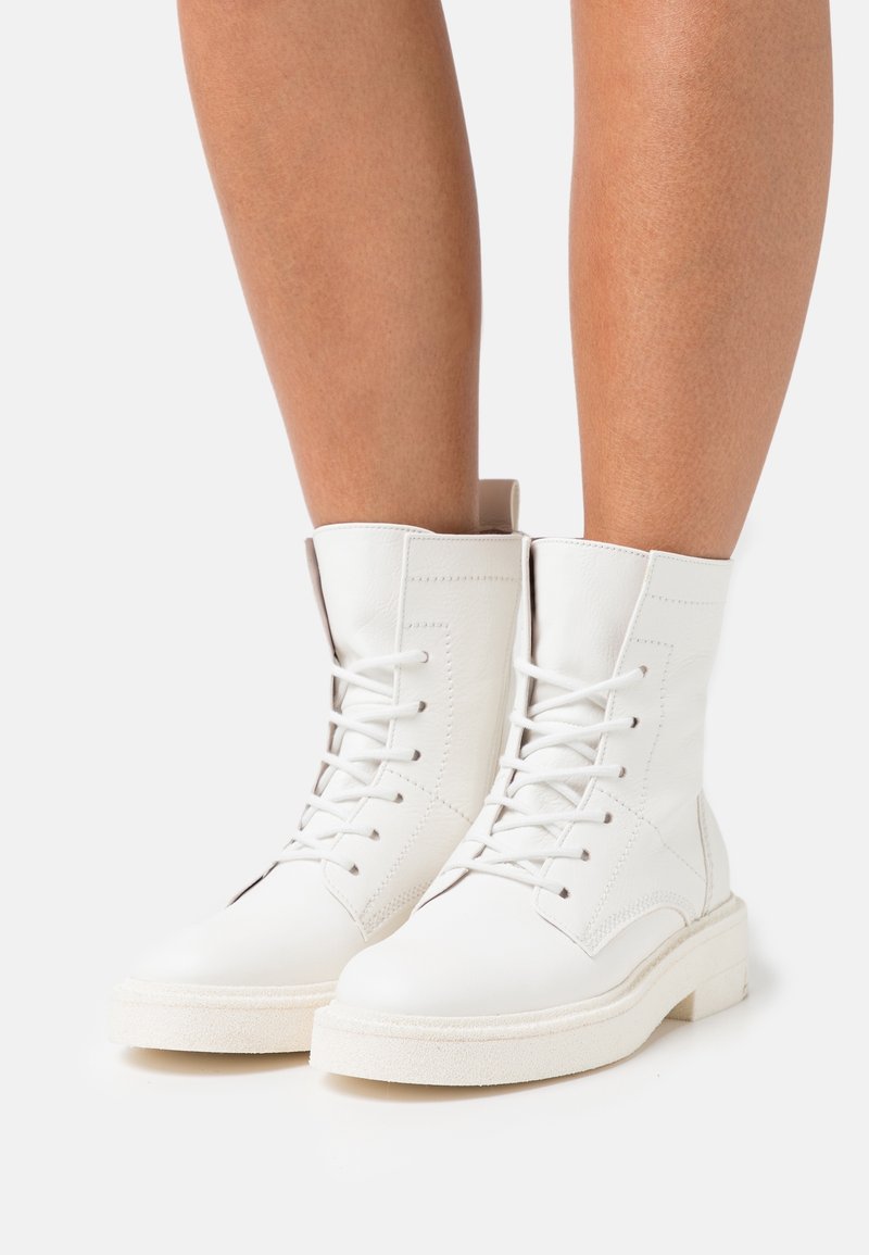 Bottes blanches à lacets montant à la cheville, en cuir texturé, avec bout rond et semelle plate à plateforme. Elles présentent des détails de couture et une boucle au talon pour faciliter l'enfilage.