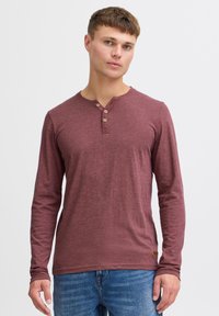 Langærmet maroon henley skjorte med tre knapper ved kraven, lavet af blødt stof, parret med blå jeans og en struktureret overflade.