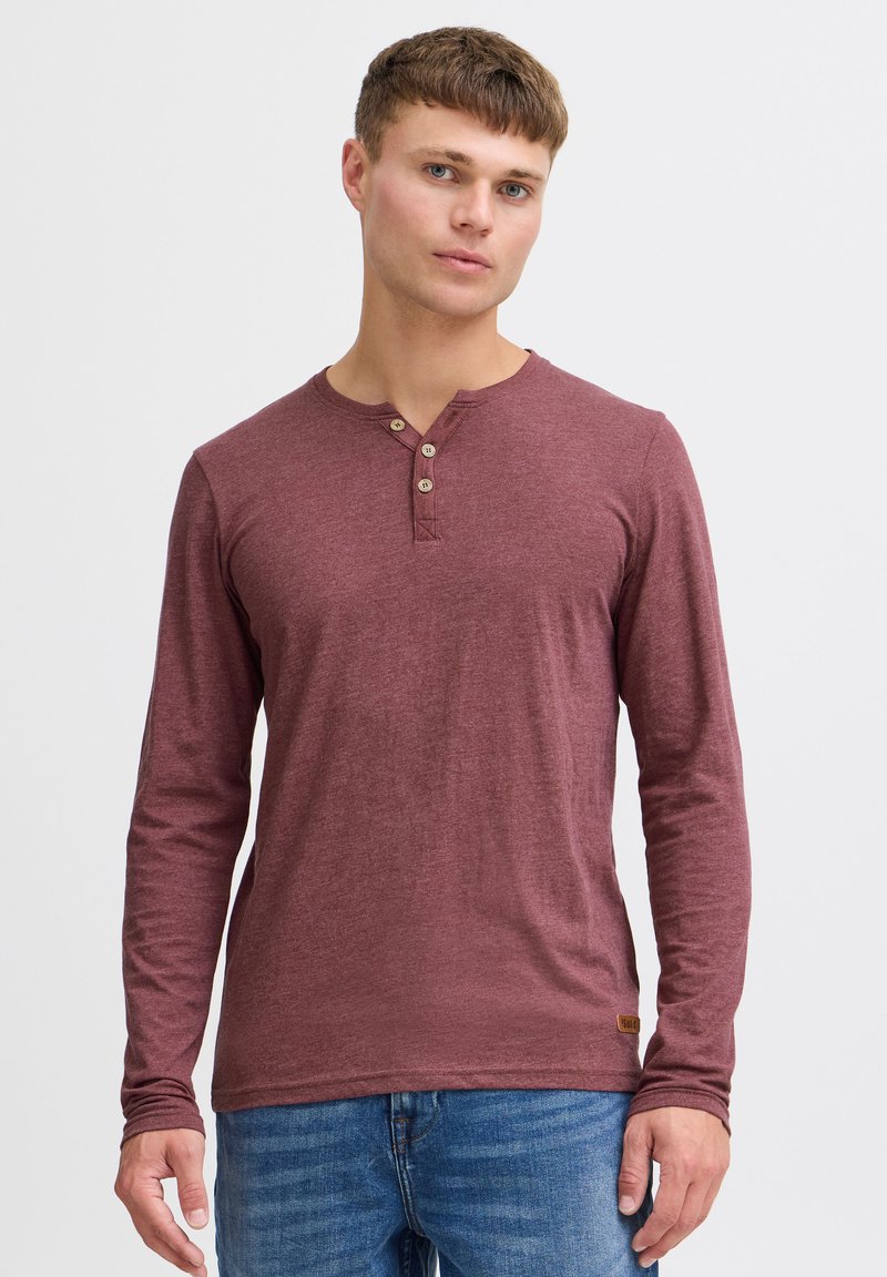 Langærmet maroon henley skjorte med tre knapper ved kraven, lavet af blødt stof, parret med blå jeans og en struktureret overflade.