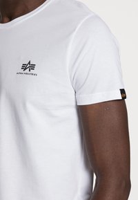 Detailní záběr osoby nosící bílé tričko s krátkým rukávem a černým logem Alpha Industries na hrudi a visačkou na rukávu.