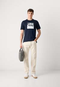 T-shirt a maniche corte blu navy con una stampa rettangolare bianca che riporta la parola "BOSS". Abbinato a pantaloni bianco sporco e una borsa grigia.