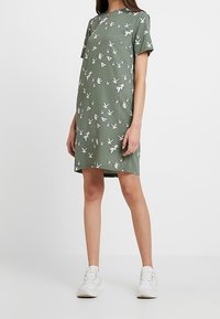 Grünes kurzärmeliges Kleid mit einem Vogelmotiv in Weiß. Gerader Schnitt mit rundem Ausschnitt. Getragen mit weißen Sneakers. Weicher Stofftextur.