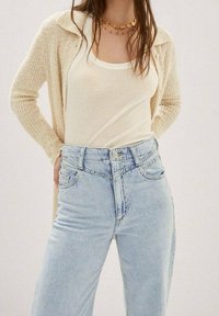 Cardigan beige en tricot porté sur un débardeur blanc, assorti à un jean en denim clair à taille haute avec légère usure au niveau des poches.