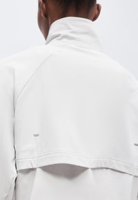 Personne portant une veste blanche à col montant vue de dos, montrant des coutures détaillées et un panneau arrière ventilé.
