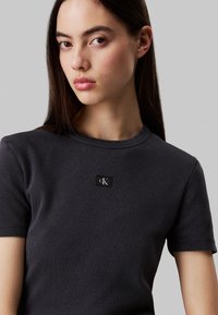 T-shirt in cotone a coste grigio scuro con design aderente, maniche corte e una piccola patch nera con logo sul petto.