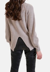 Krémový žebrovaný svetr s asymetrickým lemem, který má dlouhé rukávy a uvolněný střih, v kombinaci s černými kalhotami z umělé kůže.